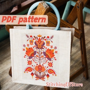 Op de afbeelding: Een beige jute tas met een blauwe handvat. De tas heeft een kleurrijk geborduurd ontwerp met een gestileerd bloemenpatroon in oranje, rood, paars en groen. De tekst "StitchingElfStore" is afgedrukt op de onderkant van de tas.