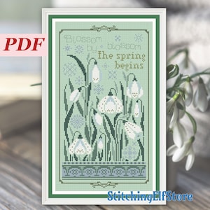 Può includere: Uno schema di ricamo a punto croce con uno sfondo verde, bucaneve bianchi e il testo "Blossom by blossom the spring begins".