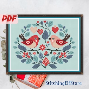 May include: Cross stitch pattern featuring two red birds with hearts on their chests, surrounded by a floral design with red and blue accents. The pattern is set against a light blue background.