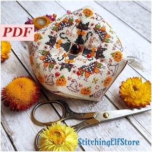 Könnte beinhalten: Ein cremefarbenes, kreuzgesticktes Nadelkissen mit Halloween-Motiven, darunter schwarze Katzen, Kürbisse und das Wort "Boo!". Das Nadelkissen ist mit Nadeln und einer dekorativen Perle verziert. Goldfarbene Scheren und Trockenblumen sind ebenfalls im Bild.