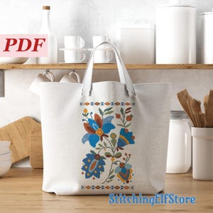 Puede incluir: Un bolso tote blanco con un diseño floral de punto de cruz en azul, naranja y marrón. El bolso tiene asas grises y el texto "StitchingElfStore" en la parte inferior. La palabra "PDF" está en la esquina superior izquierda.