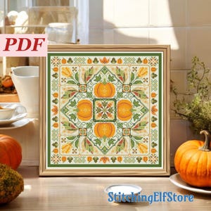 May include: A square cross stitch pattern in a wooden frame featuring a central design of four orange pumpkins surrounded by green leaves, yellow flowers, and geometric borders. The pattern includes small pumpkin icons along the edges.
