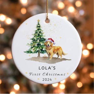 May include: White ceramic Christmas ornament with a watercolor illustration of a golden retriever wearing a Santa hat standing next to a green Christmas tree. The ornament has the text "LOLA'S First Christmas 2024" printed on it.