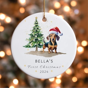 May include: White ceramic Christmas ornament featuring a dachshund wearing a Santa hat, standing next to a green Christmas tree. The ornament has the text "BELLA'S First Christmas 2024" in a decorative font, with gold accents.