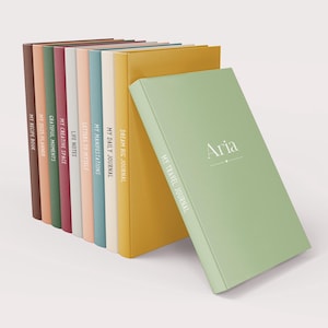 May include: A stack of ten colourful journals with different titles, including "My Daily Journal", "Dream Big Journal", "My Recipe Book", and "My Travel Journal". The top journal is green and has the name "Aria" written on the front.