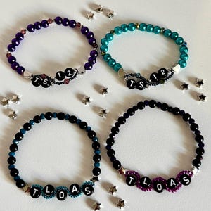 Könnte beinhalten: Vier Armbänder mit Perlen in Lila, Türkis, Schwarz und Dunkellila. Jedes Armband hat Buchstabenperlen, die Wörter und Zahlen buchstabieren. Kleine sternförmige Charms sind um die Armbänder herum verstreut.