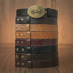May include: A stack of leather belts in various shades of brown and black. The top belt features a brass-colored buckle with a horse bit design. Some belts have metal stud details. The belts are arranged on a wooden surface.