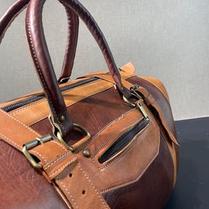 May include: A brown and tan leather duffel bag with a top handle and a shoulder strap. The bag has a zipper closure and metal hardware. The leather is stitched together with white thread.