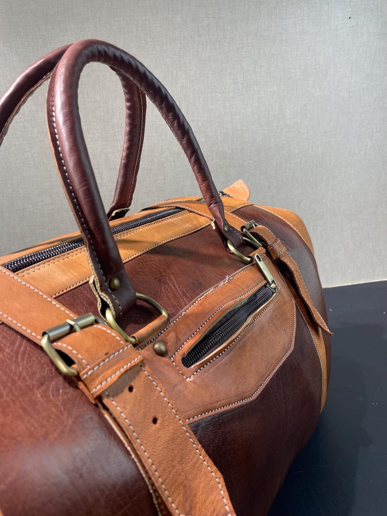 May include: A brown and tan leather duffel bag with a top handle and a shoulder strap. The bag has a zipper closure and metal hardware. The leather is stitched together with white thread.