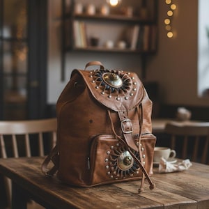 Può includere: Zaino in pelle marrone con decorazioni metalliche e borchie. Lo zaino ha una tasca frontale, spallacci regolabili e una chiusura con fibbia. È su un tavolo di legno in un caffè.