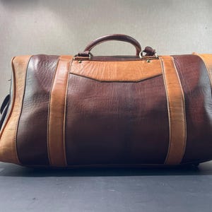 May include: A large, rectangular leather duffel bag in shades of brown and tan. The bag features a top handle and vertical straps, with a lighter brown panel on the front. The bag is designed for travel.