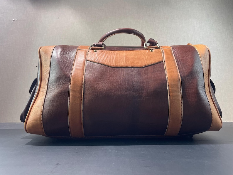 May include: A large, rectangular leather duffel bag in shades of brown and tan. The bag features a top handle and vertical straps, with a lighter brown panel on the front. The bag is designed for travel.