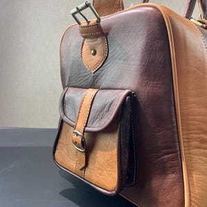 May include: A two-tone leather duffel bag in shades of brown and tan. The bag features a small front pocket with a buckle closure and a top handle. The leather has a textured appearance, and the bag appears to be well-crafted.