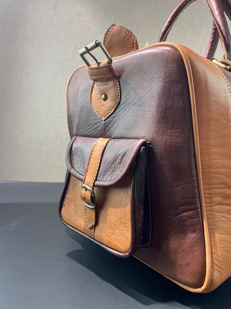 May include: A two-tone leather duffel bag in shades of brown and tan. The bag features a small front pocket with a buckle closure and a top handle. The leather has a textured appearance, and the bag appears to be well-crafted.