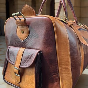 May include: A leather duffel bag in shades of brown and tan. The bag features a front pocket with a buckle closure, a top handle, and a shoulder strap. The bag is made of high-quality leather and has a classic, vintage look.