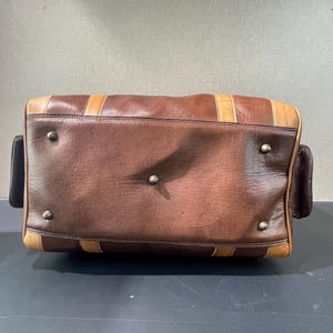 May include: A brown leather duffel bag with tan accents and metal studs. The bag has a rectangular shape with rounded corners and two small side pockets. The leather appears worn, giving it a vintage look.
