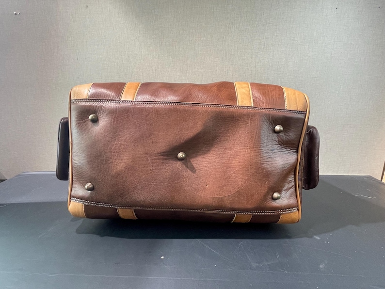 May include: A brown leather duffel bag with tan accents and metal studs. The bag has a rectangular shape with rounded corners and two small side pockets. The leather appears worn, giving it a vintage look.
