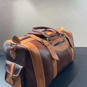 May include: A brown and tan leather duffel bag with a side pocket and top handles. The bag features a striped design with varying shades of brown and tan leather, metal hardware, and a zippered closure.