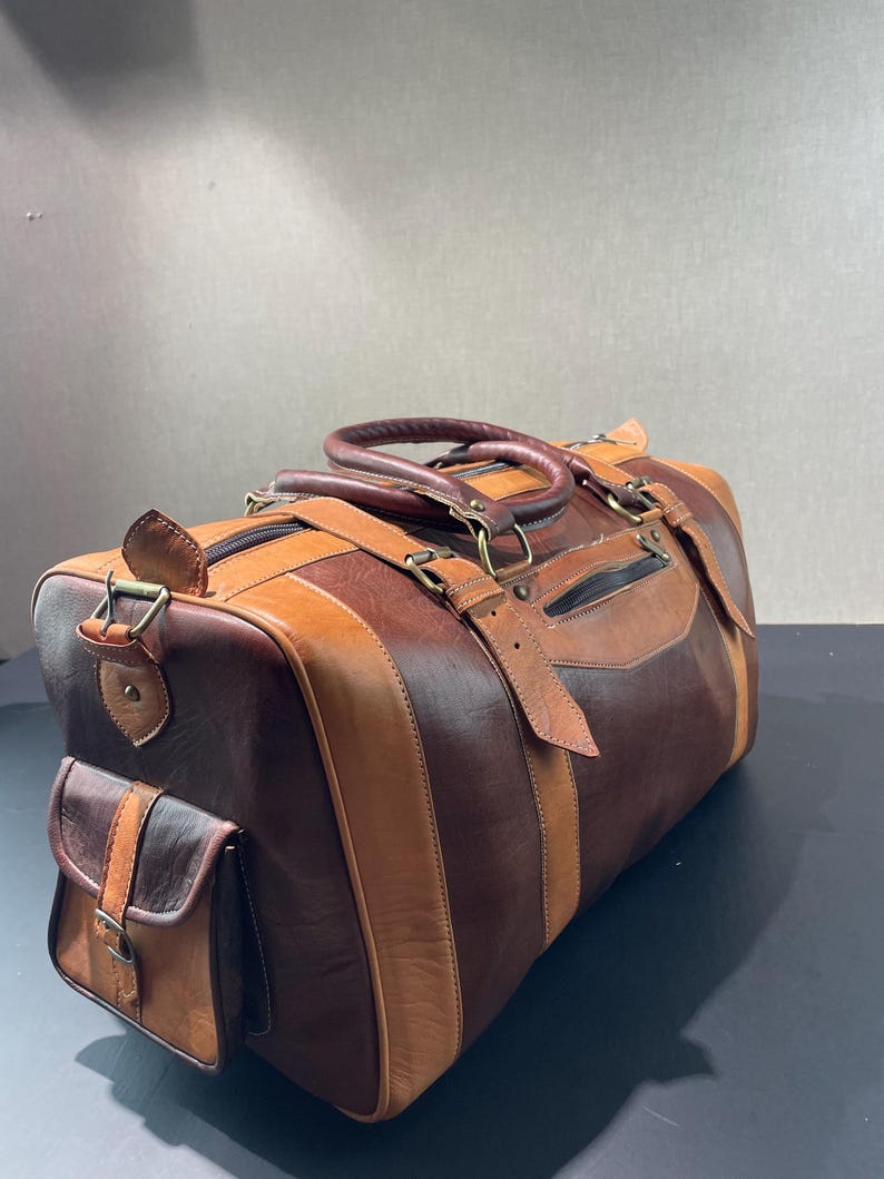 May include: A brown and tan leather duffel bag with a side pocket and top handles. The bag features a striped design with varying shades of brown and tan leather, metal hardware, and a zippered closure.
