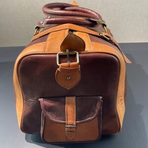 May include: A brown and tan leather duffel bag with a top handle and a front pocket. The bag has a rectangular shape with a buckle closure and a shoulder strap. The leather has a distressed look.