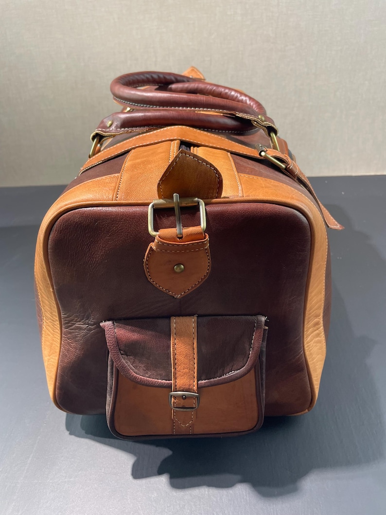 May include: A brown and tan leather duffel bag with a top handle and a front pocket. The bag has a rectangular shape with a buckle closure and a shoulder strap. The leather has a distressed look.