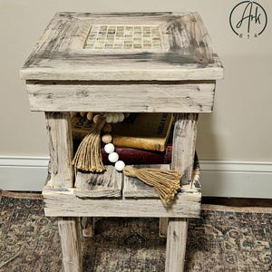 May include: A rustic, distressed wooden side table with a mosaic tile inlay on the top surface. The table features a shelf with books and decorative tassels. The design incorporates a mix of natural wood tones and a whitewashed finish.