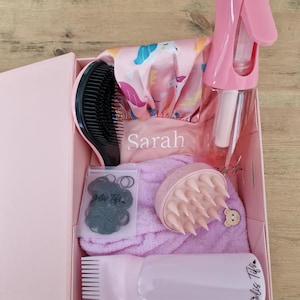 Peut inclure: Coffret cadeau rose contenant des accessoires de soins capillaires. Il comprend un vaporisateur rose, une brosse à cheveux, un bonnet de douche à motif licorne, un masseur de cuir chevelu, une serviette, des élastiques à cheveux et un peigne rose. Le nom "Sarah" est imprimé.