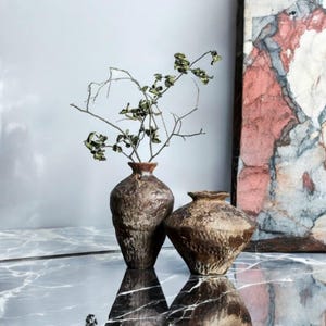 May include: Two brown ceramic vases with a rough, textured surface. The taller vase is filled with dried branches with small green leaves. The vases are sitting on a black marble surface.
