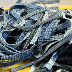 Könnte beinhalten: Ein Haufen blauer Jeansstoffstreifen, einige mit ausgefransten Kanten und einige mit sichtbaren Nähten. Die Streifen sind verheddert und überlappen sich und erzeugen ein strukturiertes und abstraktes Muster.