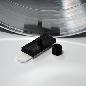 May include: A black 3D-printed record cleaner with a white base, a rectangular brush, and a small black roller. The cleaner is designed to remove dust and debris from vinyl records.