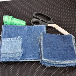 Op de afbeelding: Twee stapels blauwe denim stof vierkanten, de ene stapel is groter dan de andere. Een groene meetlint en een zilveren schaar op een zwarte stoffen achtergrond.