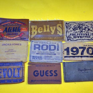 Op de afbeelding: Een verzameling rechthoekige leren jeanslabels in verschillende kleuren, waaronder bruin, beige en blauw. Elk label bevat unieke tekst en merknamen zoals "AliMk", "BellyS", "Hurley", "Jack & Jones", "Rodi", "1970", "Evolt", "Guess" en "Pepe Jeans".