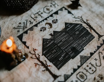 Könnte beinhalten: Ein Kreuzstichmuster mit einem schwarzen Haus und einem Halbmond darüber. Der Text "Witch" befindet sich über dem Haus und "You're" unter dem Haus. Das Muster ist auf einem beigefarbenen Stoff.