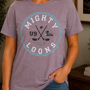 May include: A heathered purple short-sleeve t-shirt featuring a circular graphic with the text MIGHTY LOONS. The design includes two crossed hockey sticks, a hockey puck, a loon illustration, the text U9, and the words MONTBONNE CANADA.