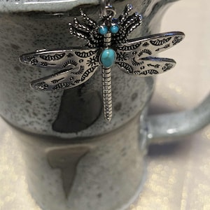 Puede incluir: Un colgante plateado con forma de libélula con detalles turquesa. La libélula tiene intrincados diseños en las alas y cuelga de una cadena. El colgante se muestra sobre una taza de cerámica moteada.