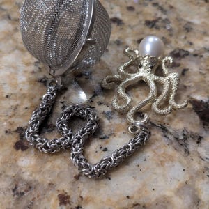 May include: A stainless steel tea infuser with a chain and a decorative octopus charm. The octopus charm is gold-colored with a pearl on top. The tea infuser is round and made of a fine mesh.