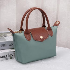 May include: A small, teal handbag with brown leather handles and a flap closure. The bag features a gold-colored button on the flap and gold-colored hardware. The handles are arched and stitched to the bag.