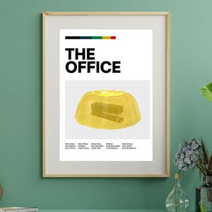 May include: Framed poster with the text "THE OFFICE" above a yellow jelly with a stapler inside. The poster is set against a teal wall. Decorative elements include hanging light bulbs and a potted plant. The poster's design is minimalist and modern.