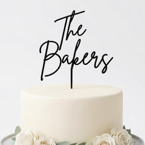 Puede incluir: Un adorno para pastel de acrílico negro con el texto The Bakers en una fuente cursiva. El adorno está insertado en la parte superior central de un pastel cilíndrico blanco liso, decorado con rosas blancas y hojas verdes en la base.