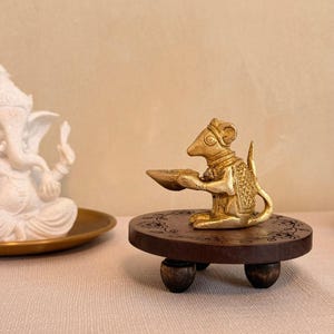 May include: A brass figurine of a mouse holding a small bowl, positioned on a round, dark brown wooden pedestal with three short legs. The mouse is depicted in a seated posture, wearing textured clothing details, and is facing toward the left side.