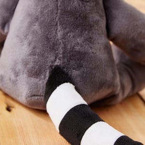 May include: A plush gray raccoon toy with a black and white striped tail. The toy is sitting on a wooden surface.