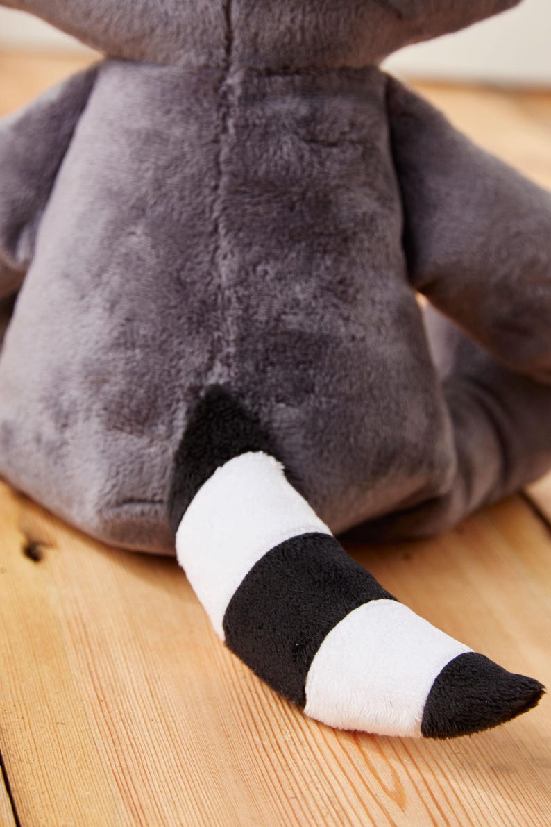May include: A plush gray raccoon toy with a black and white striped tail. The toy is sitting on a wooden surface.