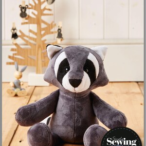 May include: A gray plush raccoon toy sitting on a wooden floor. The toy has black eyes, a black nose, and a white muzzle. The toy is wearing a black and white mask. The toy is sitting in front of a wooden tree with black and white animal ornaments. The image includes the text "Rory the Raccoon" and "Simply Sewing Patterns PDF".