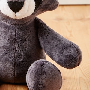 May include: A plush gray raccoon toy with black eyes and a white face. The toy is sitting on a wooden surface.