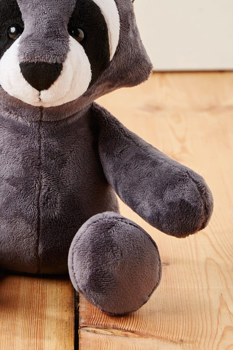 May include: A plush gray raccoon toy with black eyes and a white face. The toy is sitting on a wooden surface.