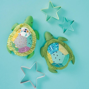 May include: Two green stuffed turtle toys with patchwork shell designs and three star-shaped cookie cutters on a light blue background. The turtles have patterned shells in various colours and textures.