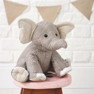 May include: A plush elephant toy with a gray and white wood grain pattern. The elephant has large ears, a long trunk, and is sitting on a pink stool. The background includes a white brick wall and tan triangle banners.