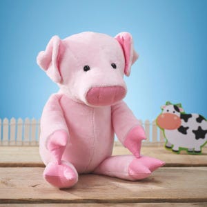 May include: A plush pink pig toy with large ears, a prominent snout, and small black eyes. The pig is sitting on a wooden surface, with a cartoon cow in the background. The toy is soft and cuddly, perfect for children.