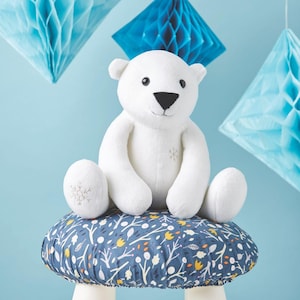 Puede incluir: Un oso polar de peluche blanco con una nariz negra y detalles de copo de nieve bordados en las patas y el pecho. El oso está sentado sobre un cojín azul y blanco estampado. Decoraciones de papel de nido de abeja azules en el fondo.