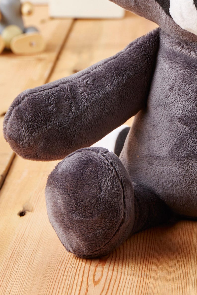 May include: A plush raccoon toy with a gray and white striped tail. The toy is sitting on a wooden surface.